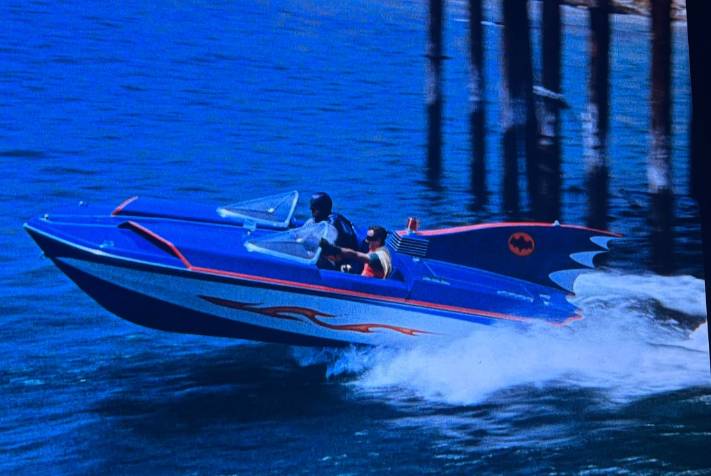 A blue and white speedboat with a bat-themed design, featuring two passengers, skimming across the water near a pier.