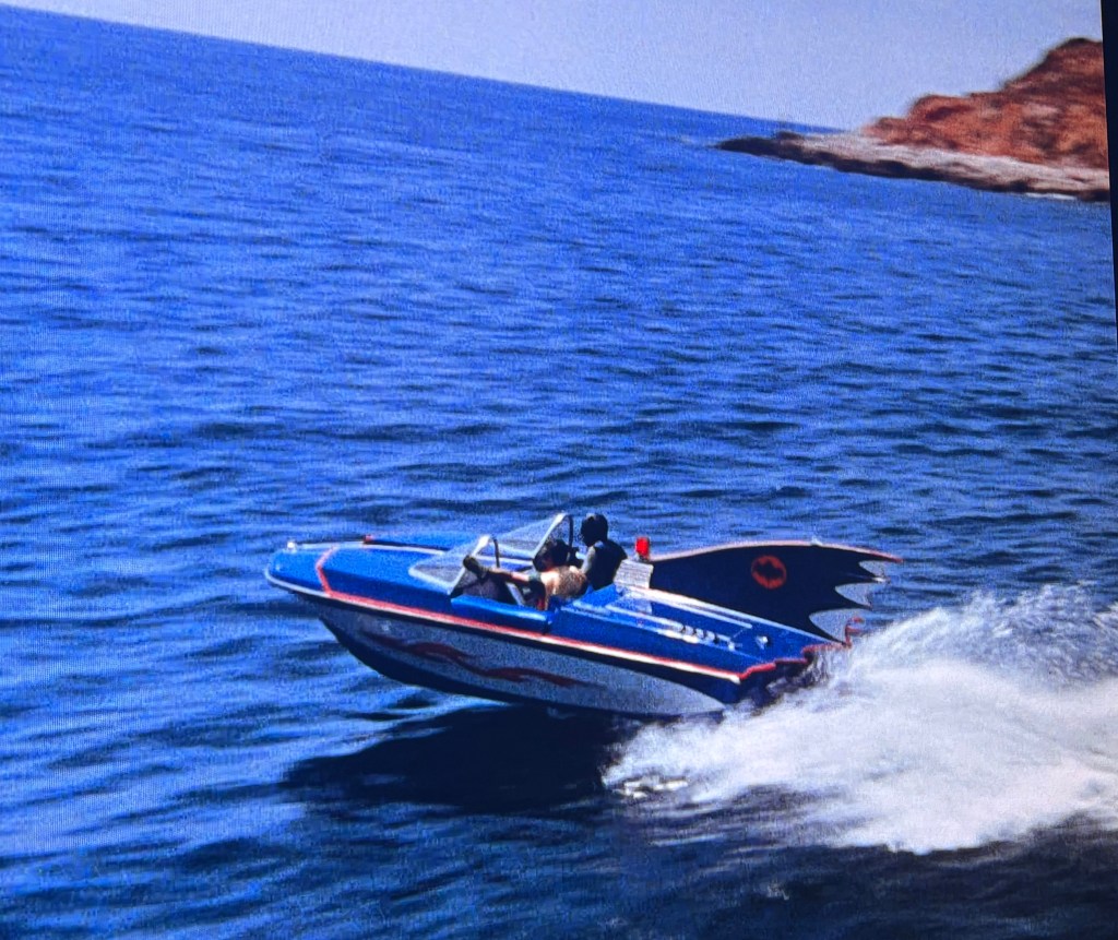 A blue speedboat with decorative red and black accents is speeding across the ocean, leaving a spray of water behind. Two people are onboard, one piloting the boat.