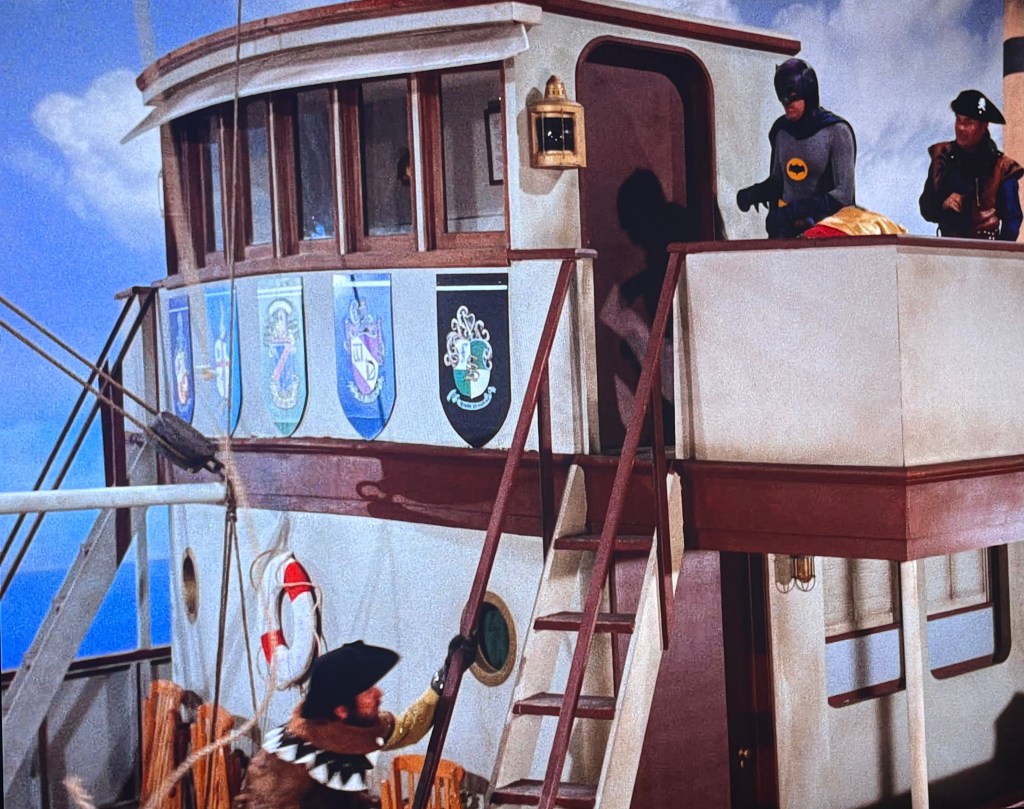 A scene featuring a ship's deck with a captain and a superhero in costume, displaying various coat of arms on the wall.