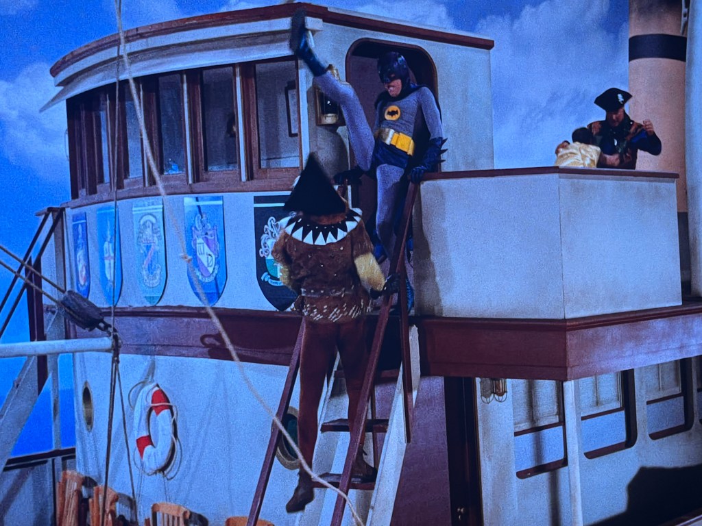 A scene from a television show featuring a superhero in a gray costume with a bat emblem, performing a kick on a pirate character wearing a hat and a colorful outfit, on the deck of a boat decorated with various shields.