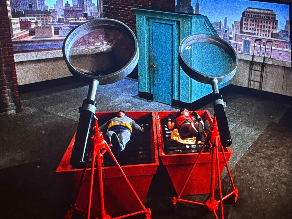 Two toy figures lie in red containers with oversized magnifying glasses positioned above them on a rooftop setting featuring a city skyline backdrop.