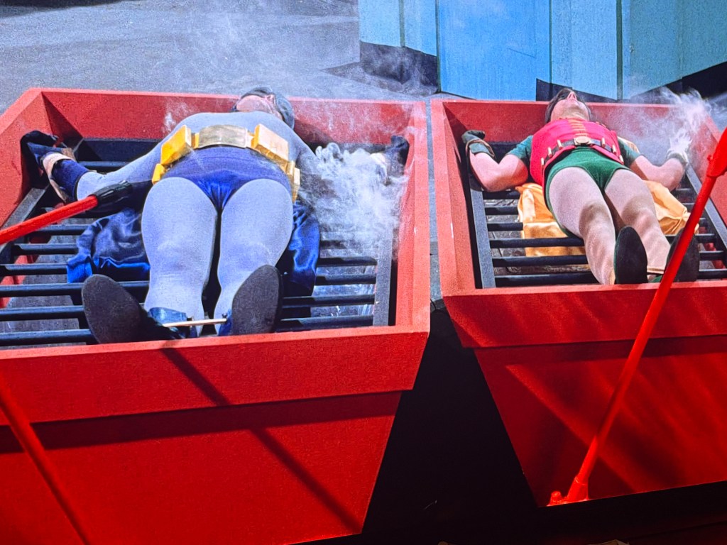 Batman and Robin lying on red devices with steam, appearing to be in distress.
