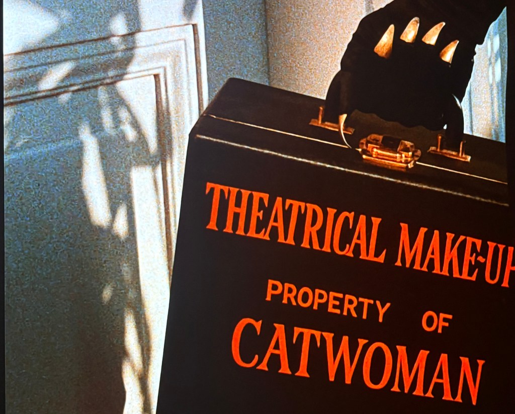 A black theatrical makeup case labeled 'PROPERTY OF CATWOMAN' with a golden handle, held by a gloved hand, casting shadows on a light-colored wall.