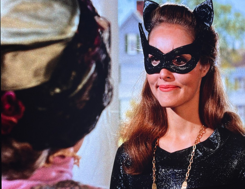A woman wearing a black cat-themed mask and a sparkling black dress, smiling at another person off-screen. She has cat ears and is accessorized with a gold chain necklace.