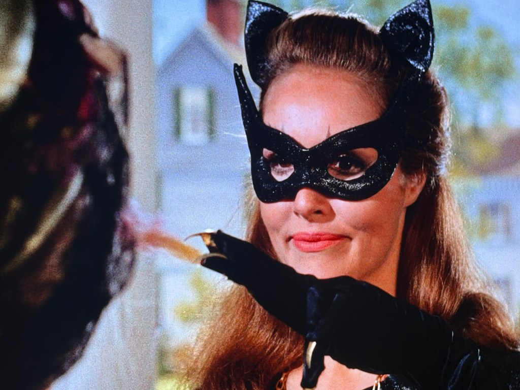 A woman wearing a black cat mask and gloves, smiling while extending her hand towards another character, with a soft-focus background of a house.