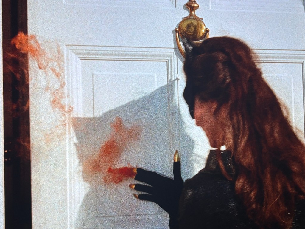 A woman in a black outfit and gloves touching a white door, leaving a red handprint, with a door knocker visible above.