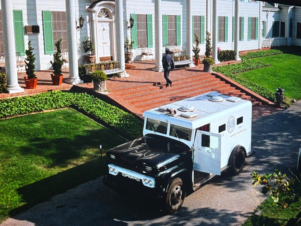 An armored transport vehicle parked in front of a large, elegant house with green shutters, featuring brick steps and landscaped gardens. A person is walking up the steps towards the entrance.