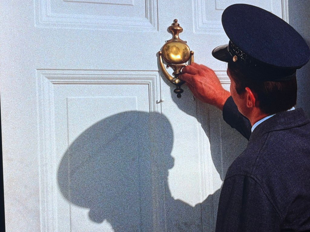 A person in a uniform knocking on a white door using a brass knocker, casting a shadow.