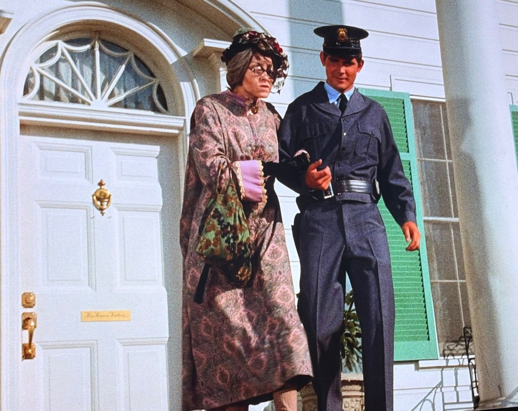 A police officer assists an elderly woman wearing a floral dress and a large hat as they descend the steps of a house, with a white door and green shutters in the background.