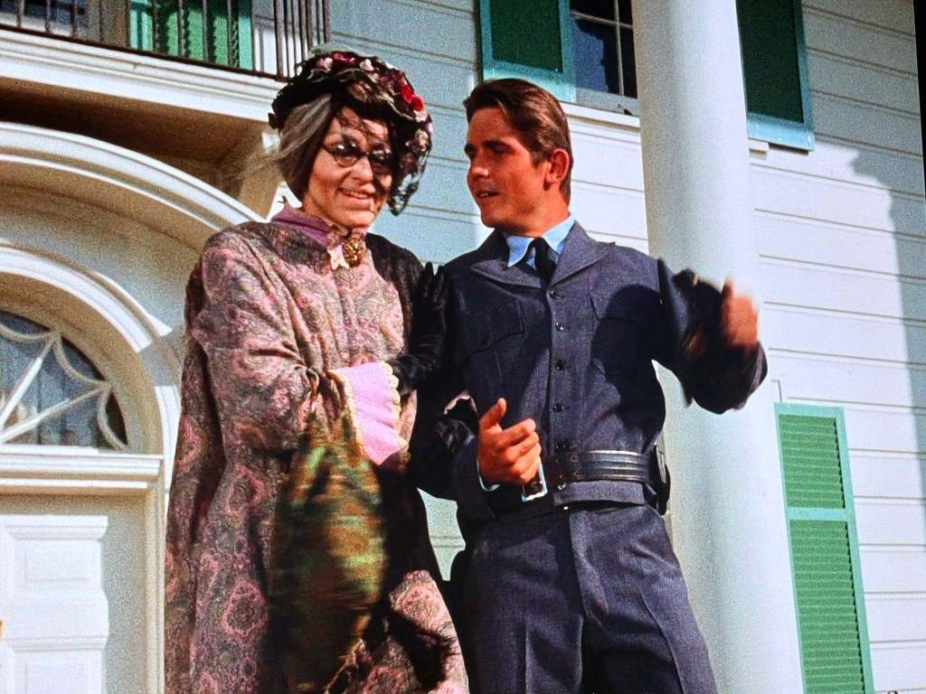 A man in a blue uniform stands smiling next to an elderly woman in a colorful, ornate dress and floral headpiece, with a grand white building featuring green shutters in the background.