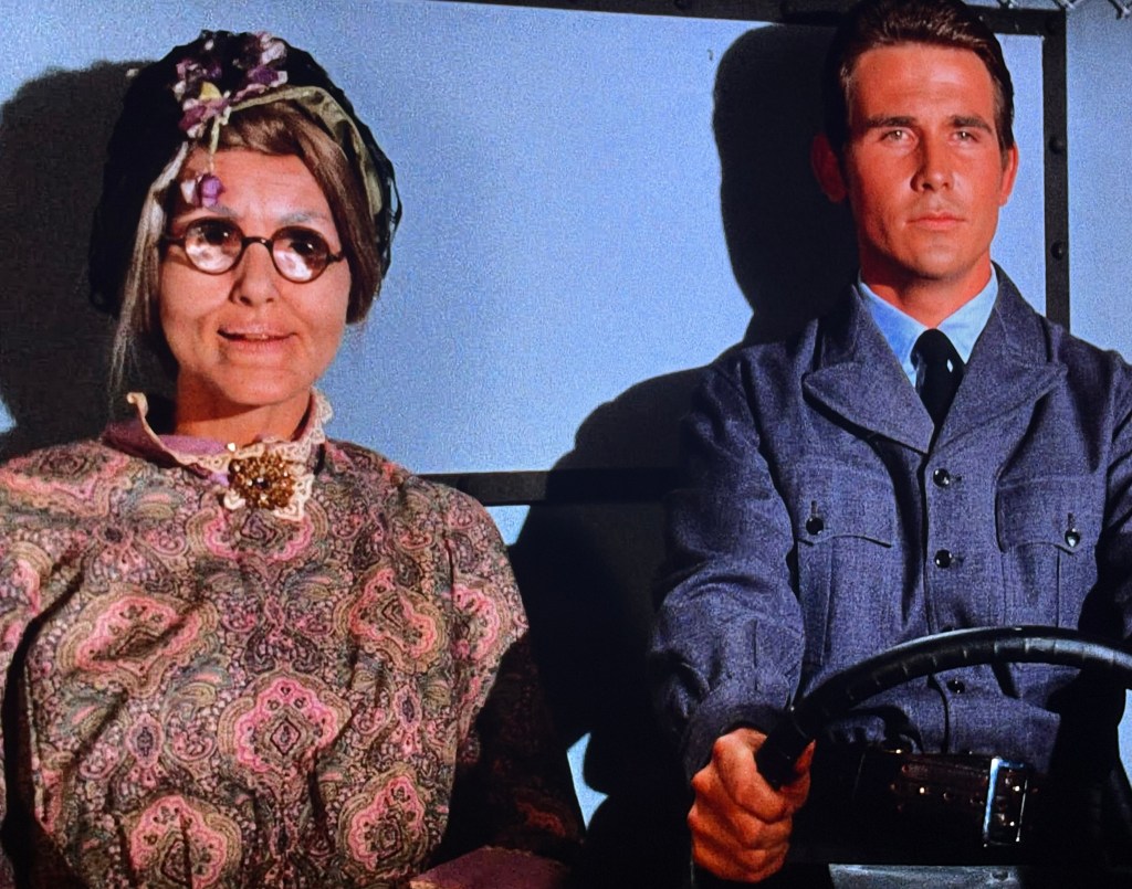 A woman dressed in an old-fashioned outfit with large glasses and a headscarf sits beside a man in a formal suit, who is driving a vehicle. The setting appears to be inside a vehicle, with the focus on their expressions.
