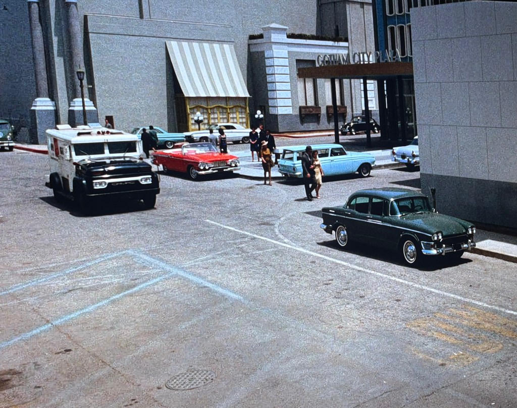 A busy street scene featuring an armored truck, several classic cars, and pedestrians walking near a building labeled Gotham City Plaza.