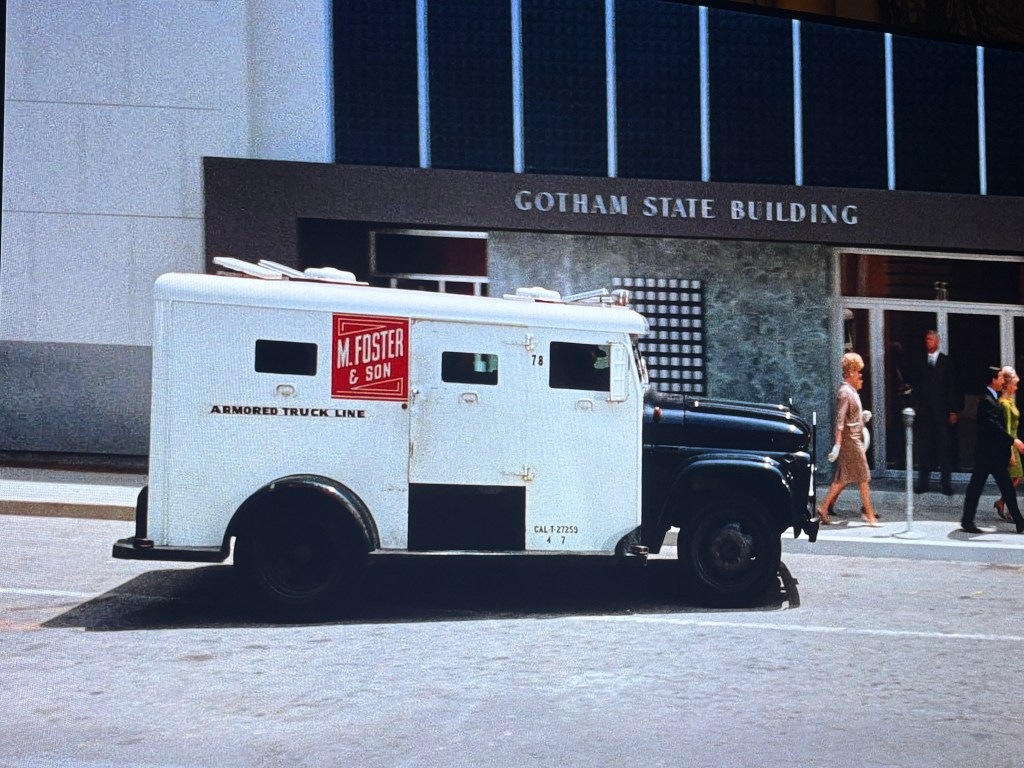 An armored truck with the sign 'M. Foster & Son Armored Truck Line' parked in front of the Gotham State Building, with pedestrians walking by on the sidewalk.