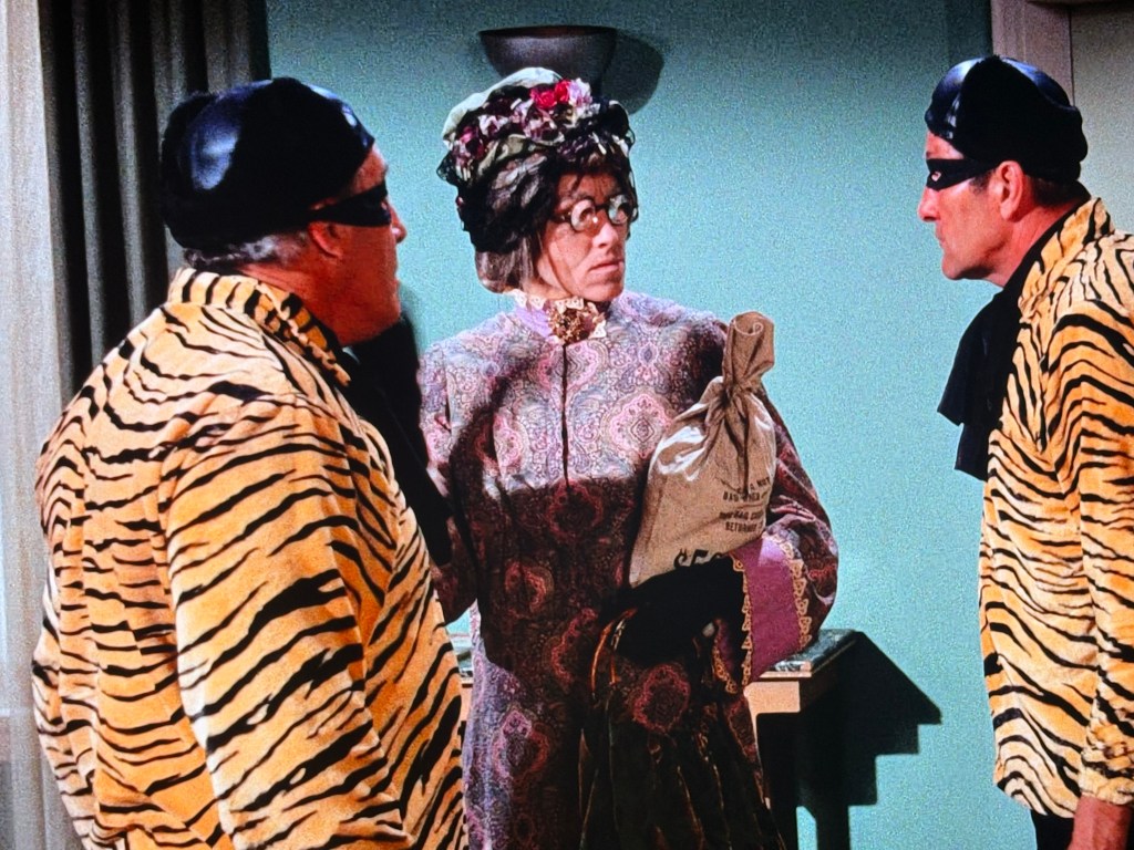 Two men in striped tiger outfits and masks confront a woman in a floral dress holding a bag.
