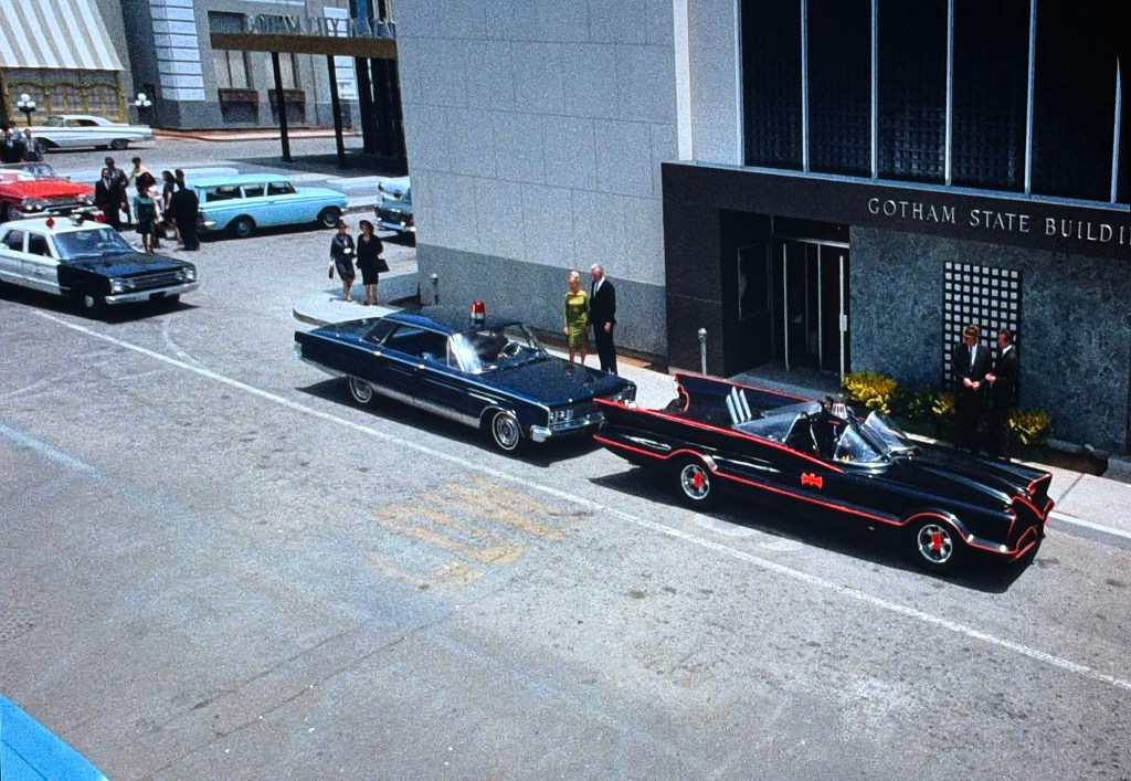 A street scene in front of the Gotham State Building featuring several vintage cars, including a black Batmobile, a police car, and other classic vehicles. A group of people is gathered nearby, dressed in formal attire.