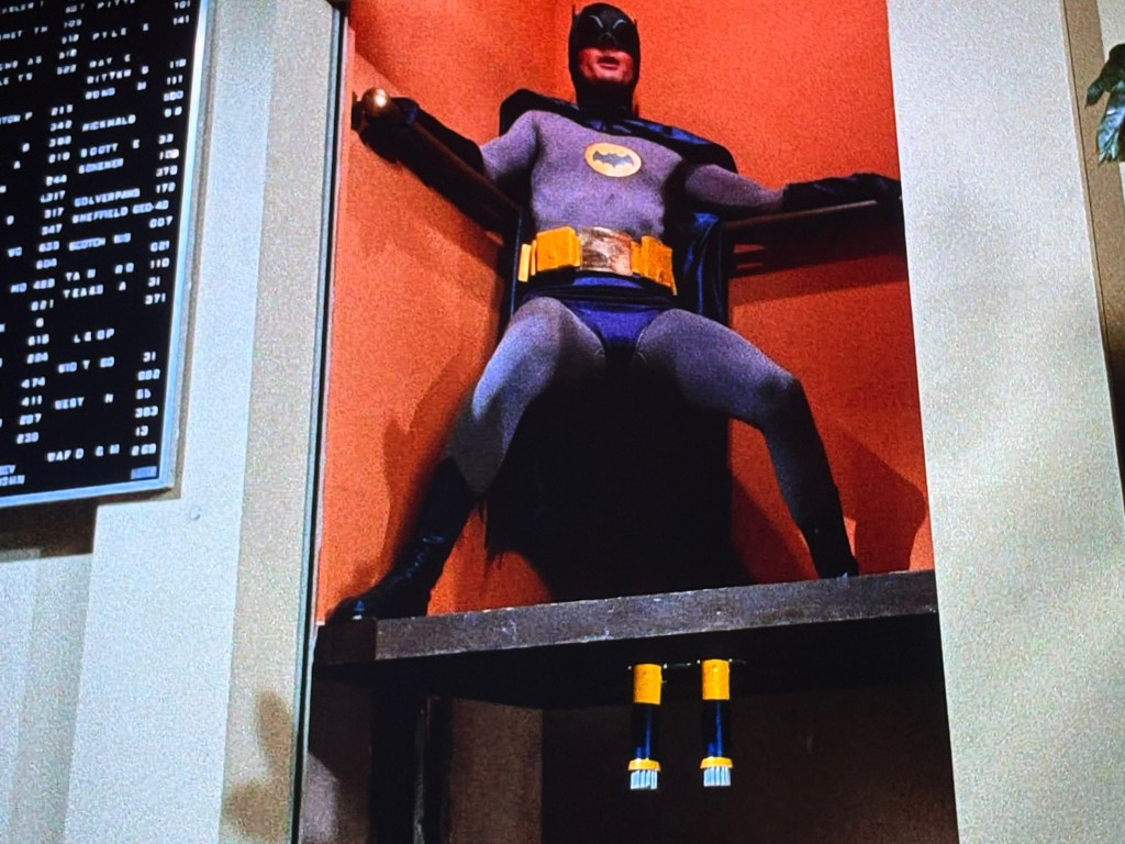 A person dressed as Batman, hanging in a posed position on an elevated platform, with a background featuring a wall-mounted display board.
