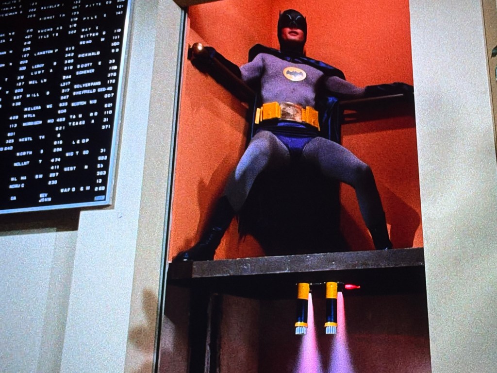A model of a superhero in a bat-themed costume, posed dramatically with arms extended, placed inside a compartment with jetpack-like devices underneath.
