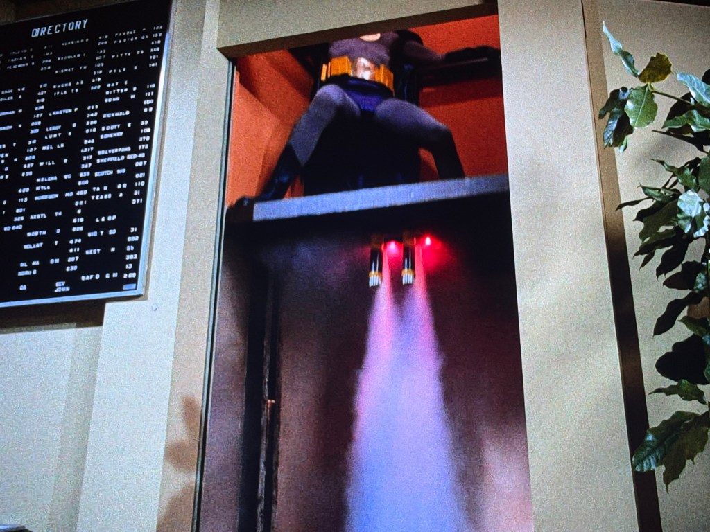 A superhero in a costume is ascending in an elevator with jet-powered rockets, emitting red flames from the back, beside a directory board.