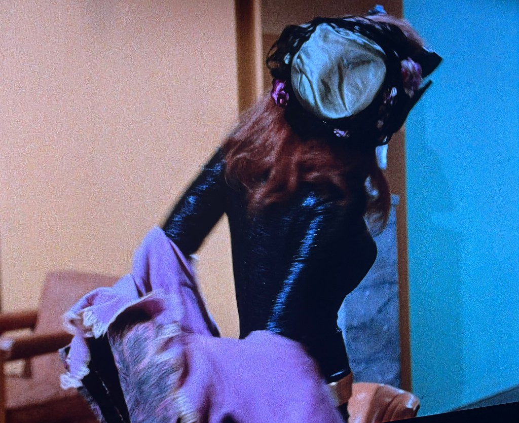 A woman in a black outfit with long red hair, wearing a decorative hat, is turning away while holding a purple shawl.