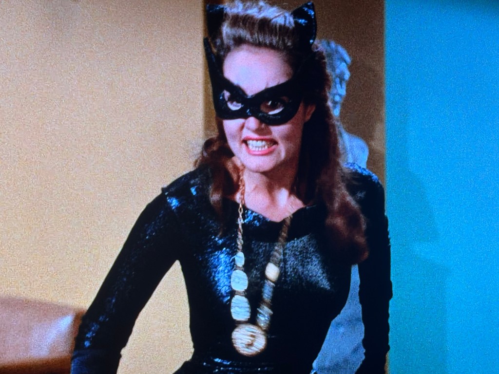 A woman dressed as a cat-themed villain wearing a black costume and a mask, with a fierce expression, standing in an indoor setting.