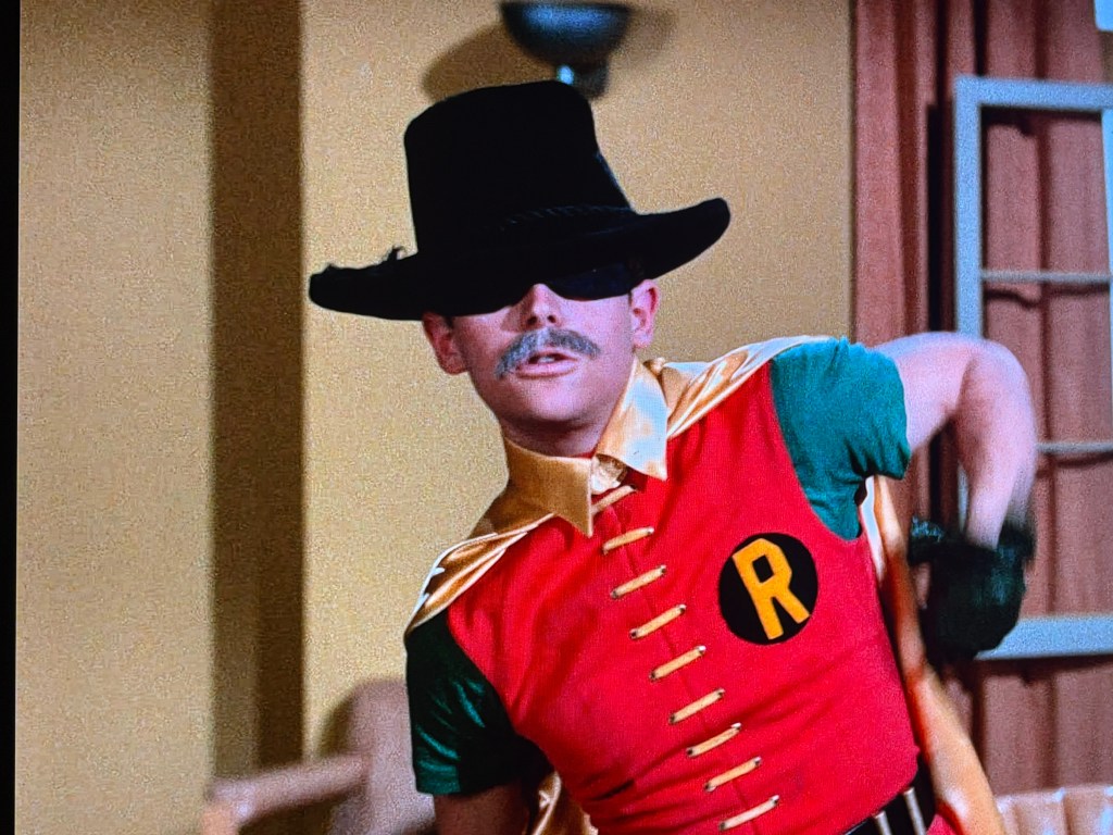 A person dressed as a superhero wearing a colorful costume with a red and green design, a large black hat, and a mask, striking a dynamic pose.