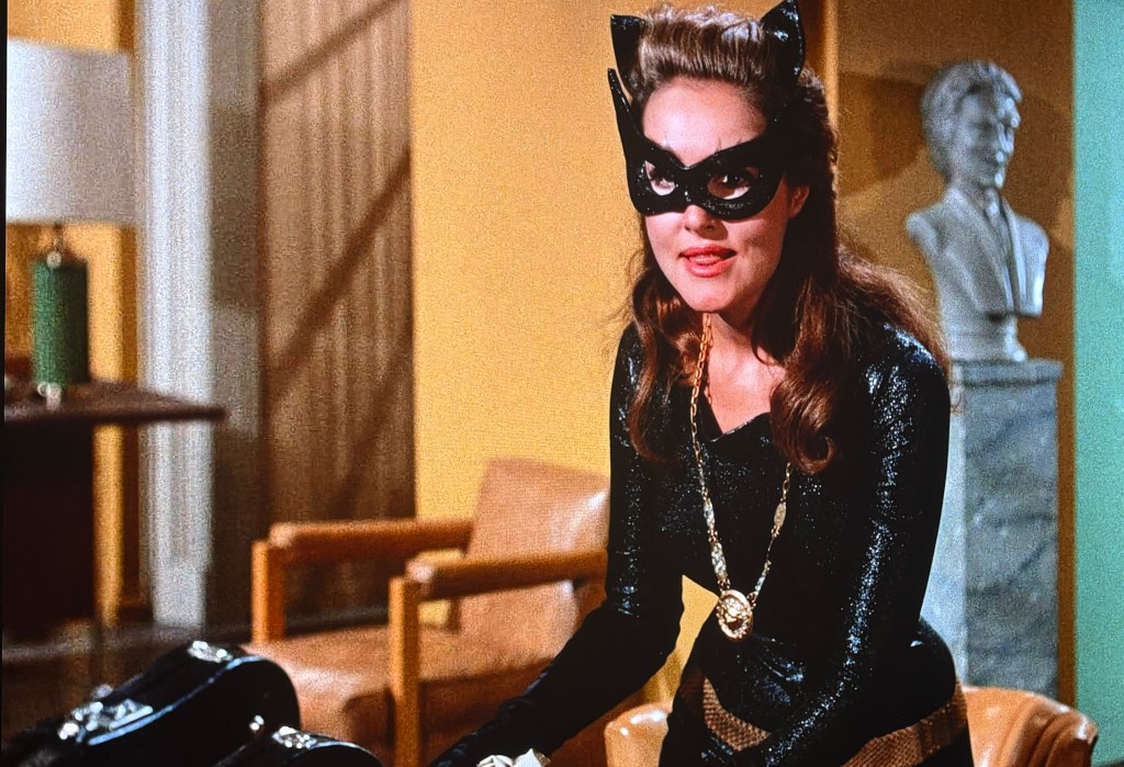 A woman dressed as a character in a black catsuit with a mask and cat ears, standing in a room with a modern interior and a statue in the background.