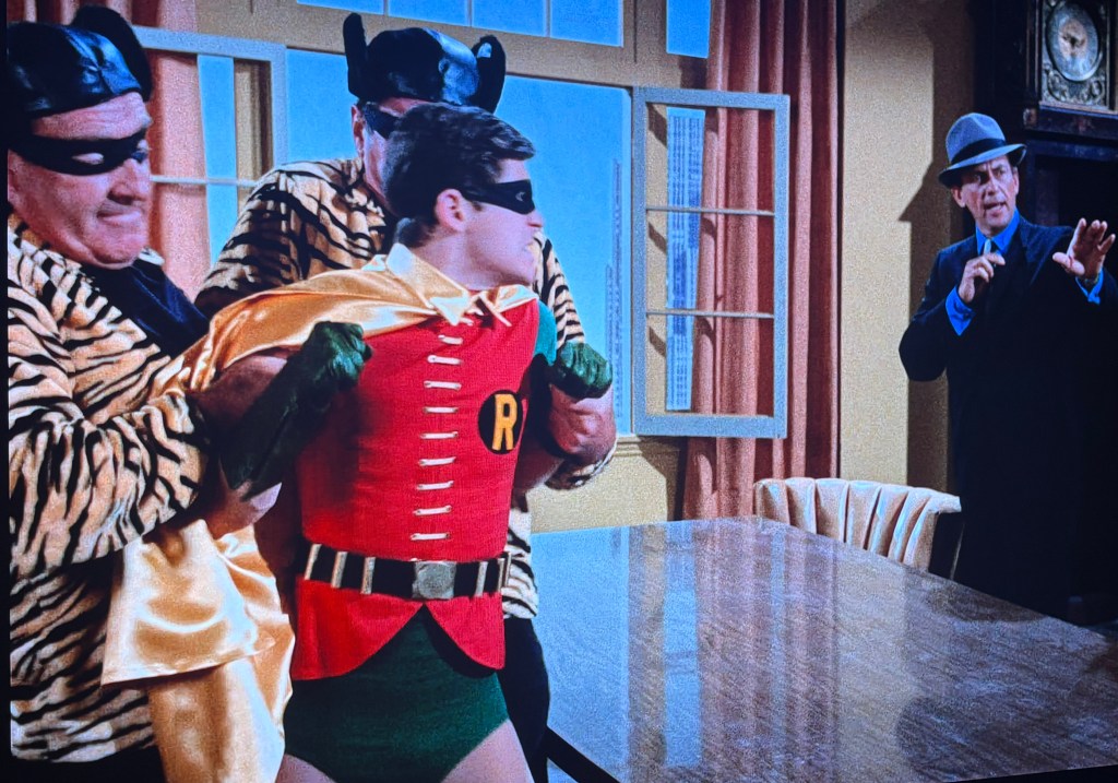 A scene depicting a superhero sidekick being restrained by two villains in tiger-striped outfits while a third villain in a suit gestures from a distance.