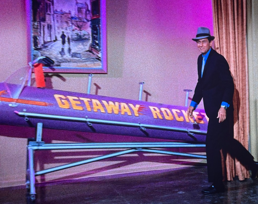 A man in a suit and hat stands beside a purple rocket labeled 'GETAWAY ROCKETS' in a brightly lit room with curtains. A painting is visible in the background.