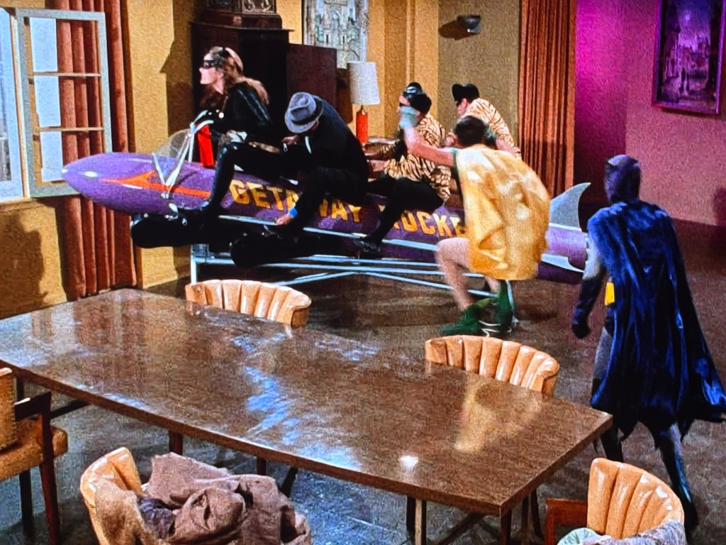 A scene depicting a chaotic moment with characters dressed in costumes, including a figure in a bat-like suit, interacting around a large table. One character is on a purple getaway rocket, while others in humorous outfits seem to be reacting to the situation.