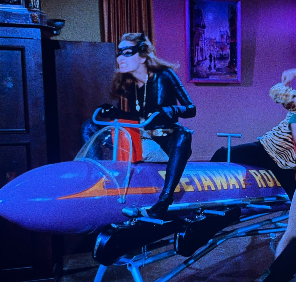 A woman in a black catsuit and mask is dramatically riding a colorful toy vehicle labeled 'Getaway Rod' indoors, while another person in a striped outfit watches.