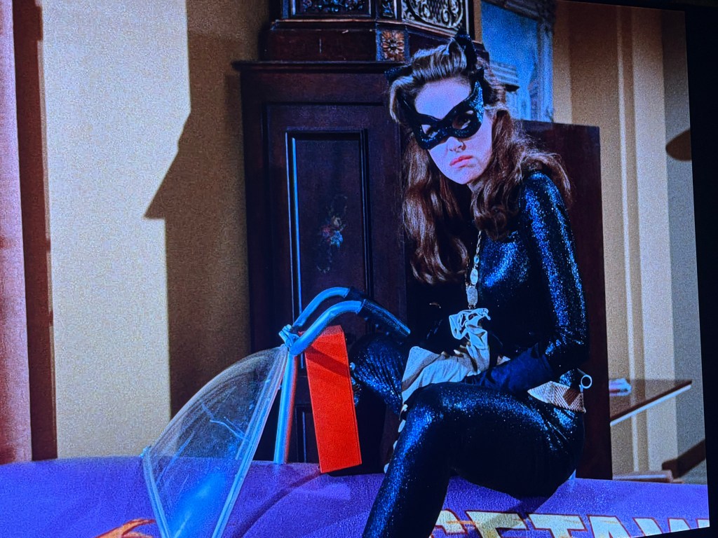 A woman dressed in a black cat costume with a mask, sitting on a purple vehicle, looking thoughtfully.