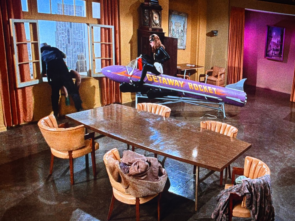 A scene featuring a stylish room with a large table and multiple chairs. A person dressed in black is climbing out of a window, while another person is inside, near a colorful rocket labeled 'Getaway Rocket' positioned in the room.