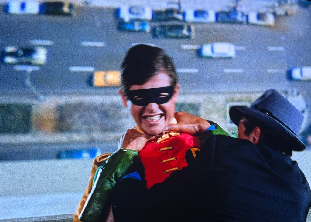 A scene depicting a young superhero in a colorful costume, featuring a red top and green accents, being confronted by a villain in a gray hat. The background shows a city street with cars below.