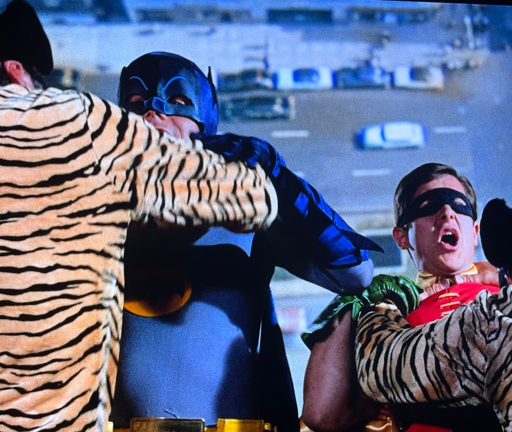 Batman and Robin being restrained by a villain in a tiger-striped costume, set against an urban backdrop.