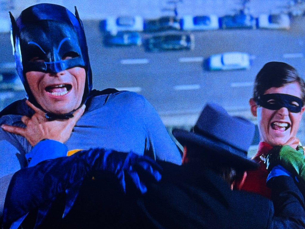 Batman and Robin struggle against a villain in a dramatic scene, with Batman appearing to choke while Robin reacts in fear.