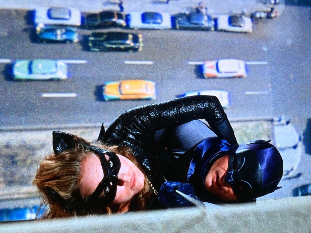 A close-up view of a female character dressed in a black cat suit and mask, looking down from a tall building, while a male character in a dark suit and mask is assisting her. Cars and a busy street can be seen below.