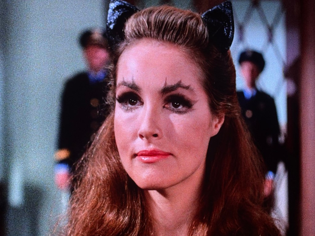 Close-up of a woman with long, wavy hair styled in a glamorous look, wearing cat-ear hair accessories and dramatic makeup, including bold eyebrows and thick eyeliner.