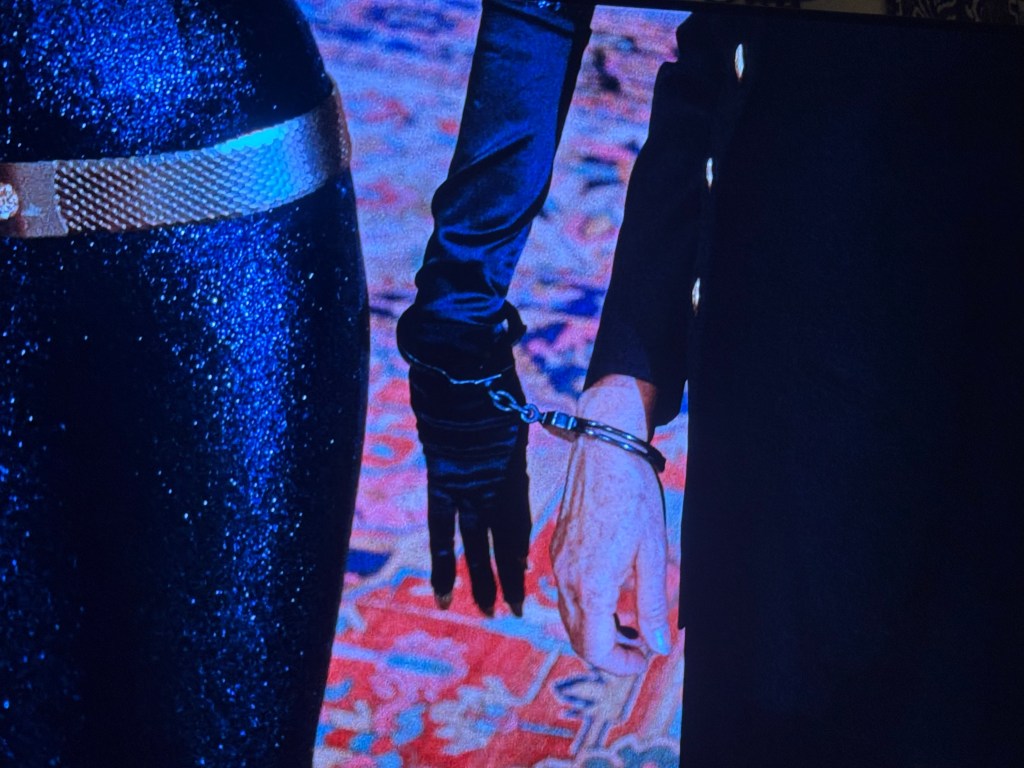 Close-up of two hands, one wearing a black glove and the other hand cuffed in handcuffs, set against a colorful patterned background.