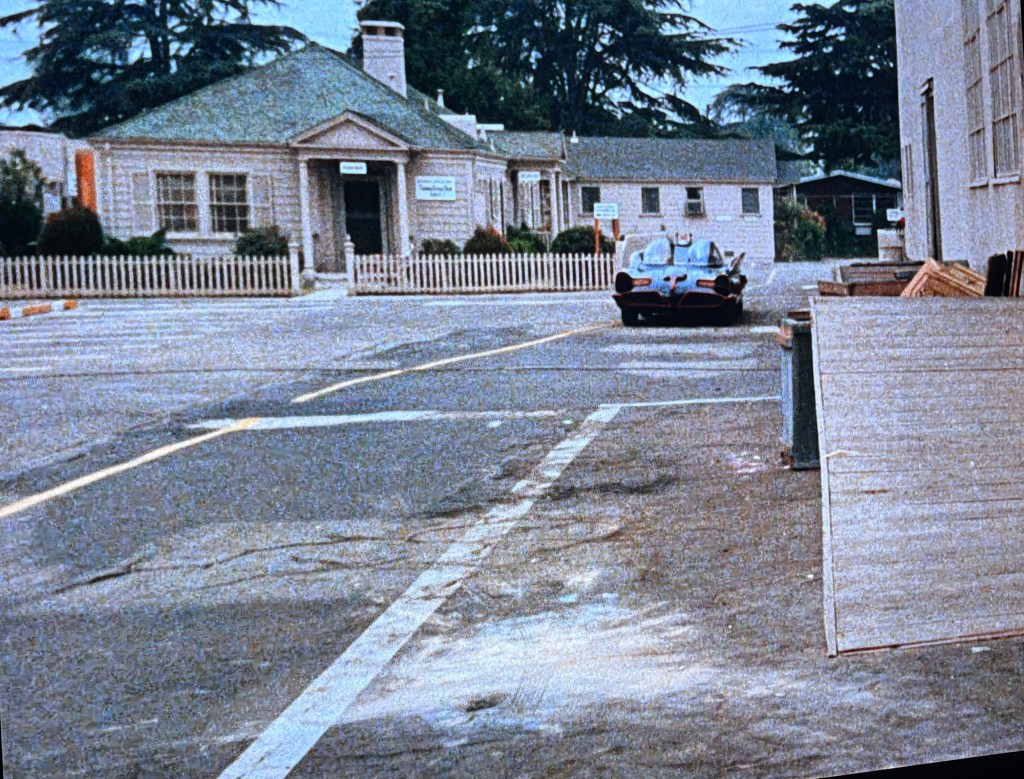 A classic car parked in an empty lot near a small building with a green roof and white fence, set against a backdrop of trees under an overcast sky.