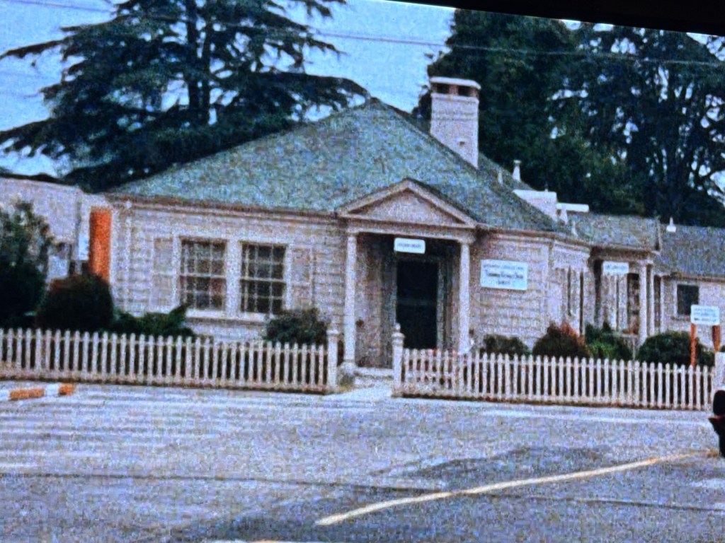 A single-story building with a green roof, white exterior, and a fenced yard. It has multiple windows and is surrounded by trees and shrubs.