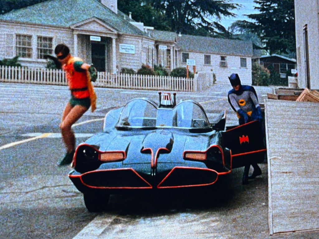 Batman and Robin beside the Batmobile in a city setting, with Batman opening the car door and Robin jumping out.