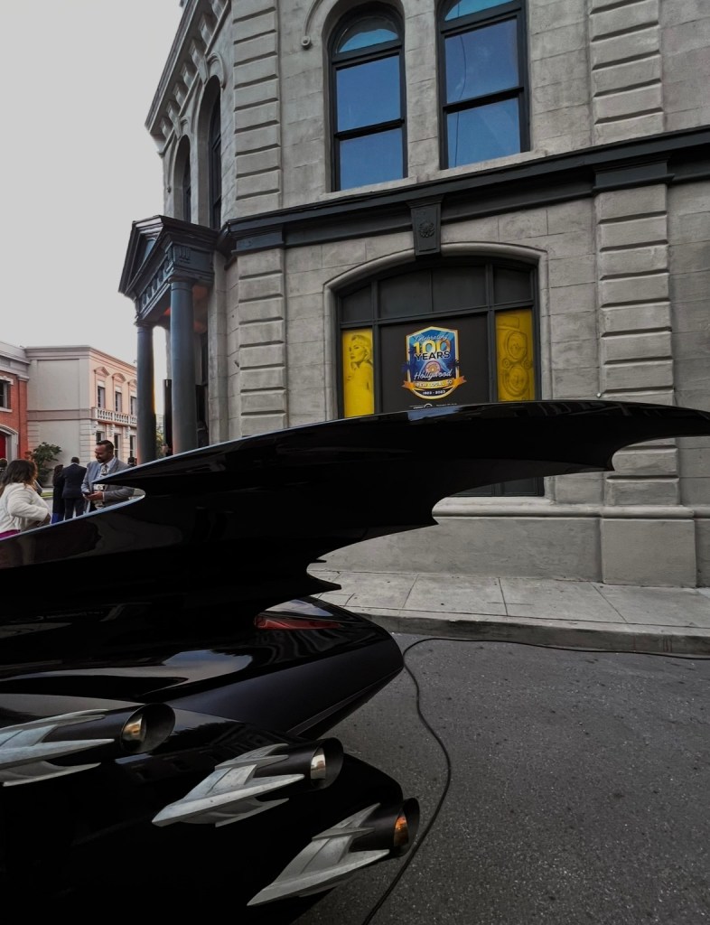 A close-up view of a black vehicle with distinct wing-like features parked on the street, with a historic building and a 100 Years of Hollywood sign in the background.