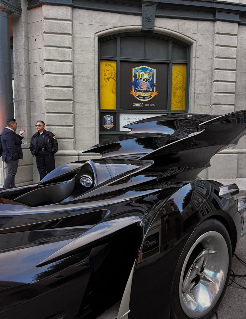A sleek black car with distinctive features is parked prominently in front of a building displaying a poster that celebrates 100 years of Hollywood. Two men are engaged in conversation nearby.