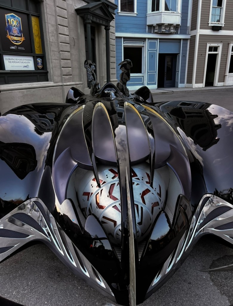 A close-up view of a sleek, futuristic black car with distinctive design features resembling a superhero motif, positioned on a street near colorful buildings.