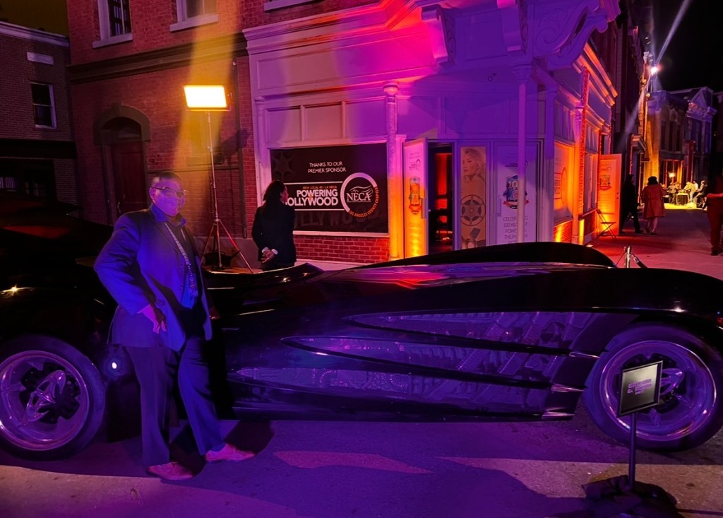 A man in a suit stands next to a sleek black car at an outdoor event. The background features decorative lighting and a sign thanking sponsors, with a vintage street setting.