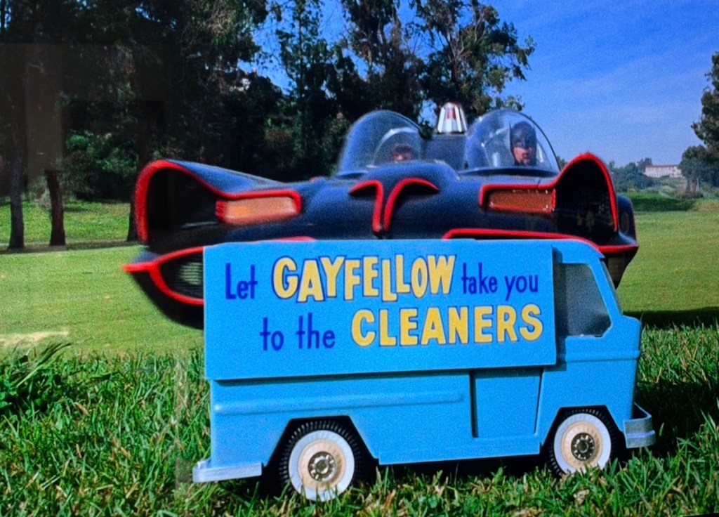 A humorous scene featuring a small blue vehicle with a sign that reads 'Let GAYFELLOW take you to the CLEANERS' in front of a sleek black car, with a grassy background.