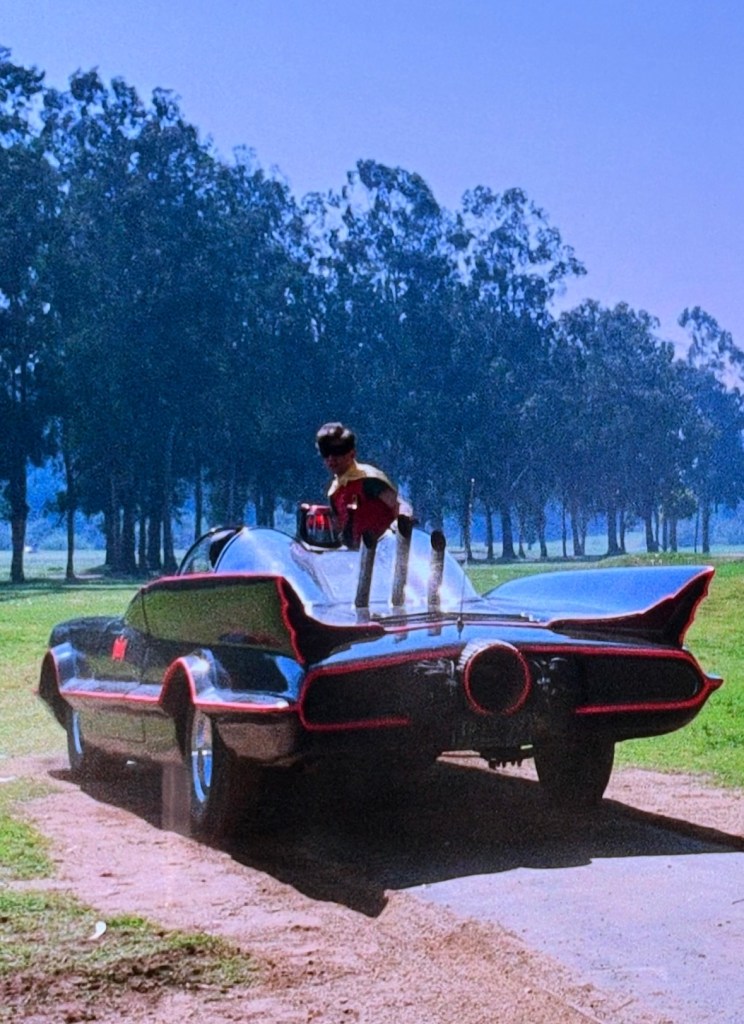 A person in a superhero costume stands atop a classic black car with distinctive fins, surrounded by green trees and grass.
