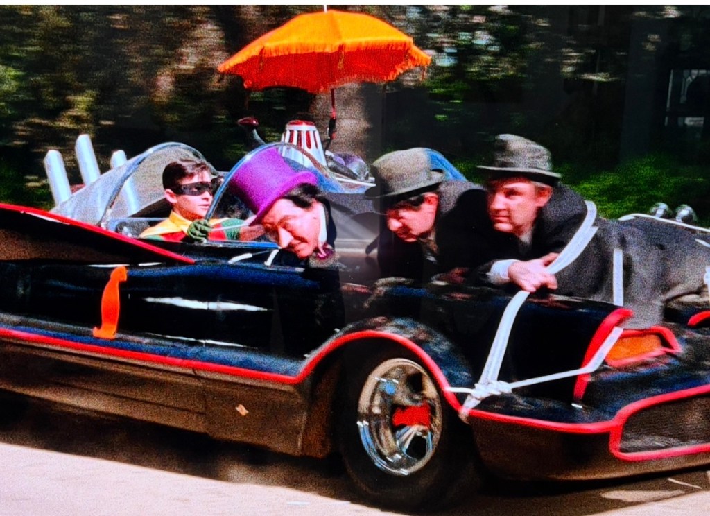 A colorful, vintage-style car with characters in costumes, including a person with a purple top hat, driving on a road with trees in the background.