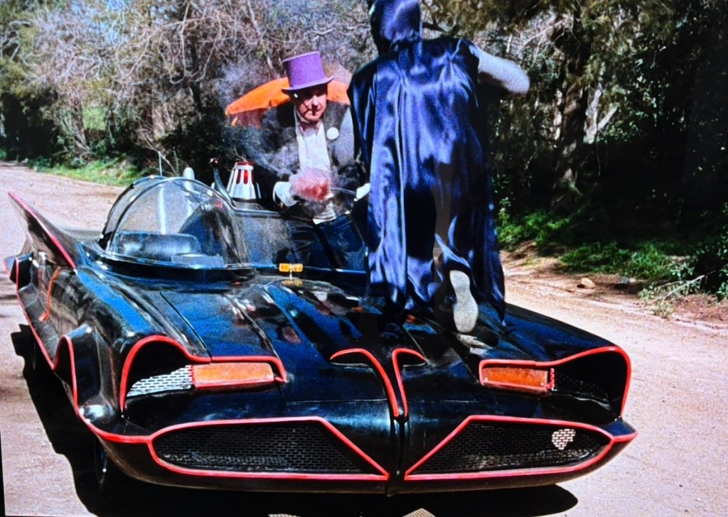 A black and red futuristic vehicle parked on a dirt road, with a character in a purple top hat and suit standing beside it, while another character in a dark cape leaps onto the car.