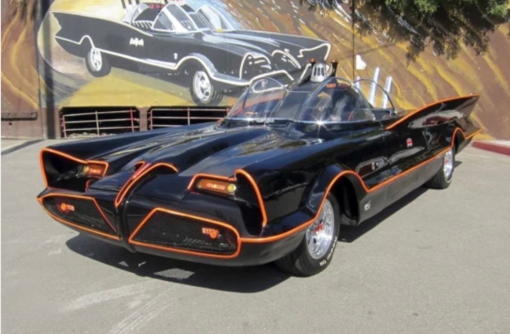 A classic black car inspired by the Batmobile, featuring distinct curves, orange accents, and a futuristic design. The background includes a mural of a similar vehicle.
